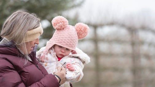Woman carrying young child in a Garden, both wearing hats and gloves.